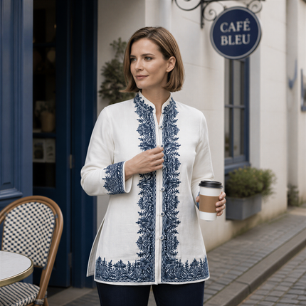 A woman wearing a white blouse with blue embroidery stands outside a cafe, holding a coffee cup.